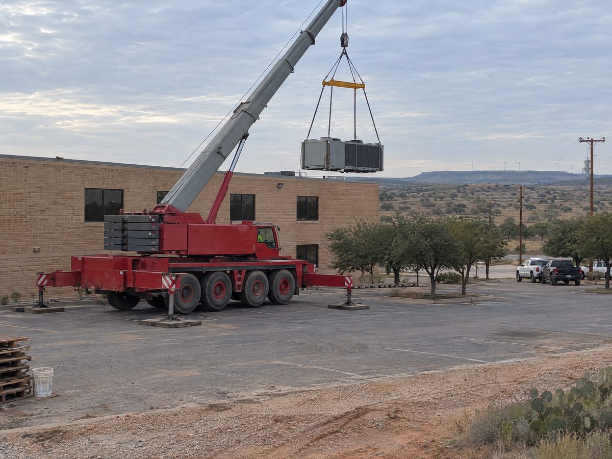 A red all-terrain crane in a parking lot lifts a rooftop chiller on a spreader bar toward the roof of a two-story tan brick building.