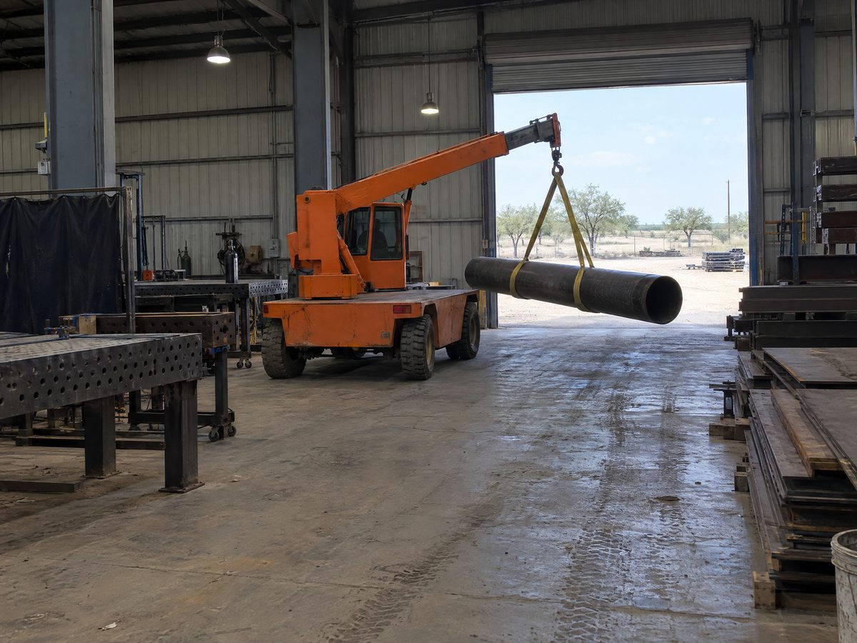 An orange carry deck crane carries a section of black steel pipe on a yellow sling down an aisle of a metal fabrication shop.