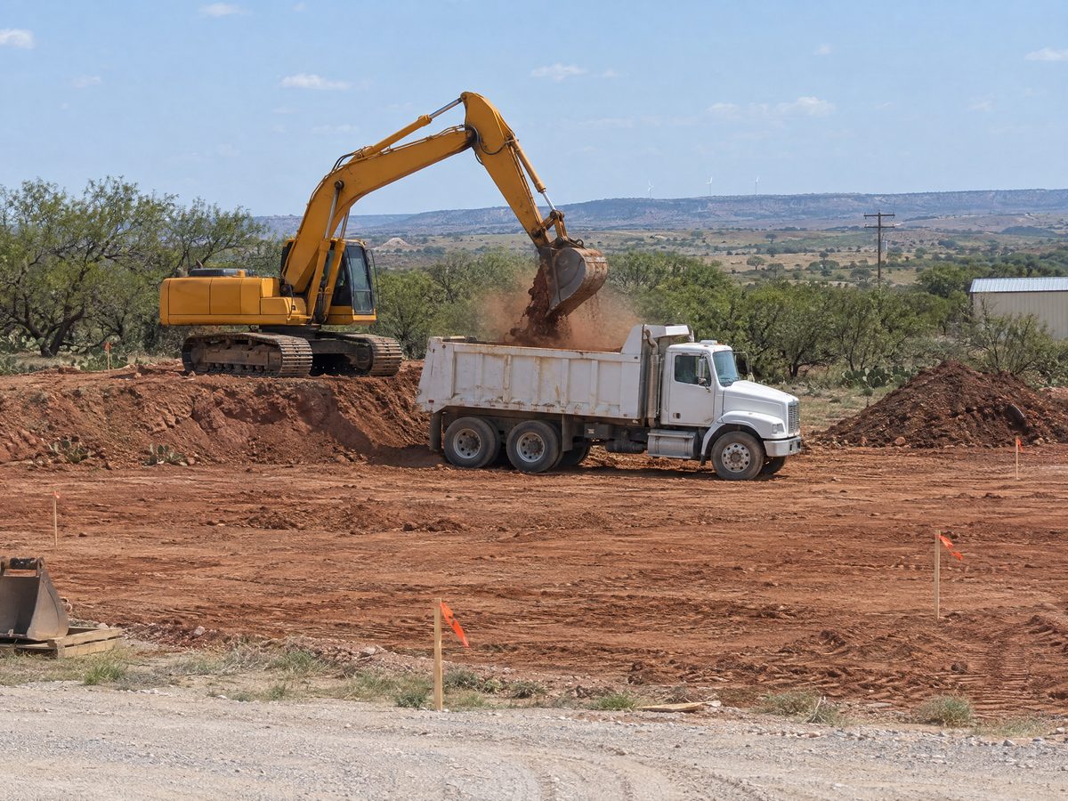 A yellow excavator on a dirt bench swings a bucket of red clay into a white dump truck on a staked building site.