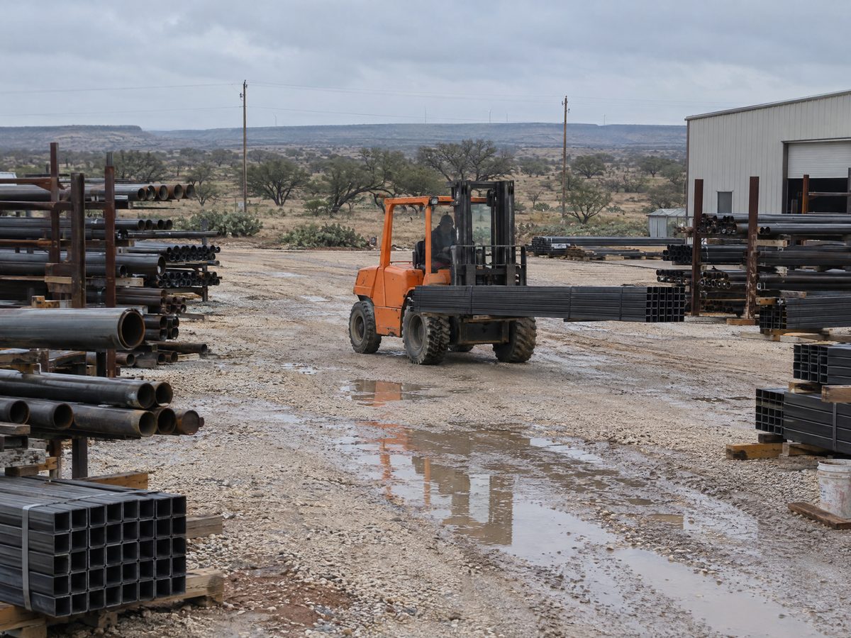 An orange pneumatic-tire forklift carries a bundle of square steel tubing between pipe racks in a wet gravel yard.
