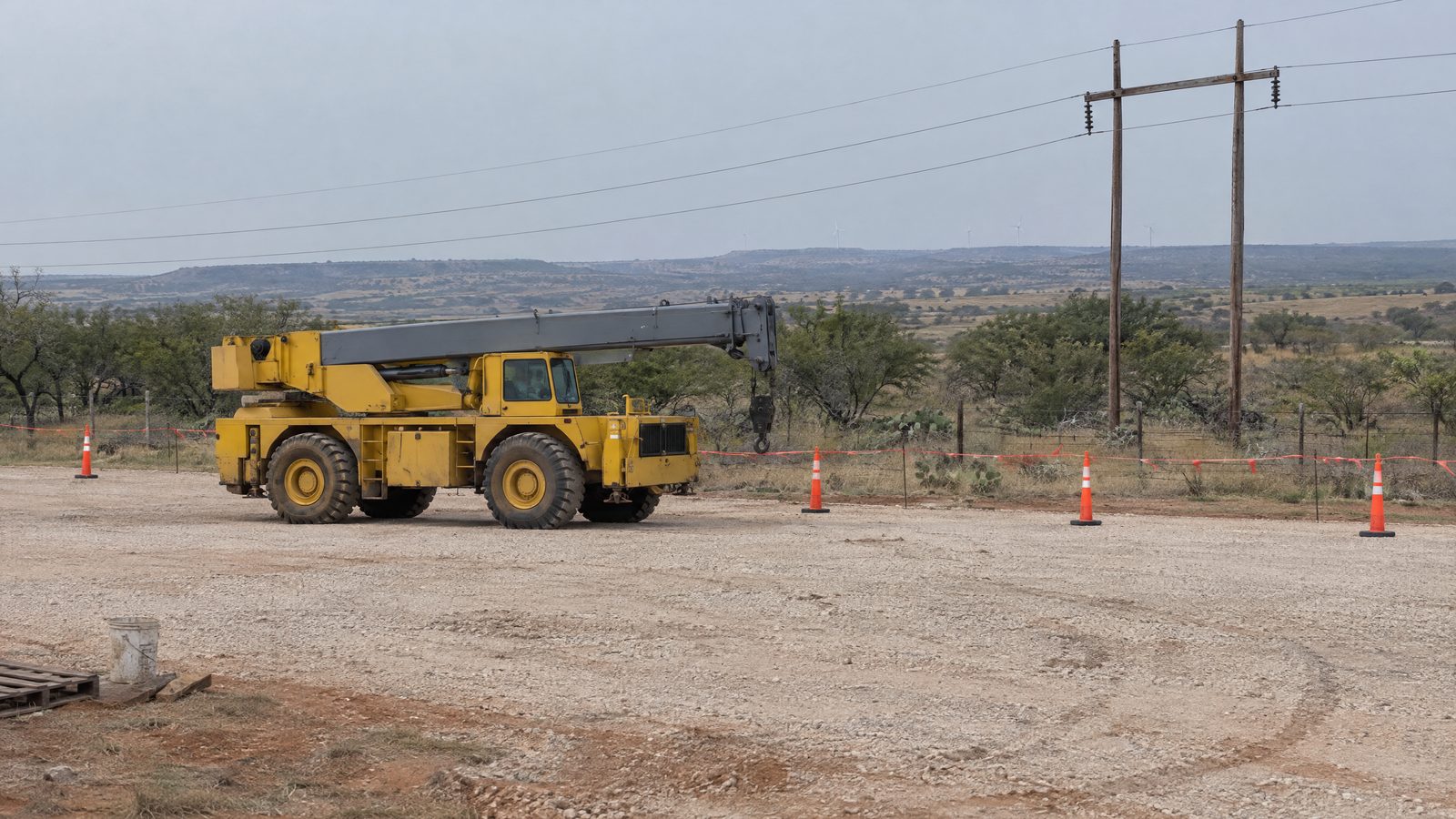 A yellow rough-terrain crane with its boom stowed sits on a caliche pad behind cones and red flagging tape, with power lines on wooden poles beyond.