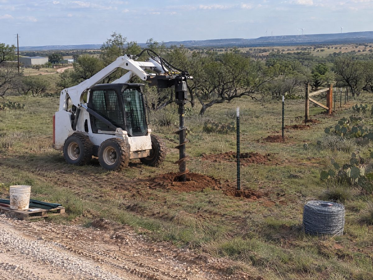 A white skid steer with an auger attachment drills post holes along a new fence line of steel T-posts in mesquite pasture.