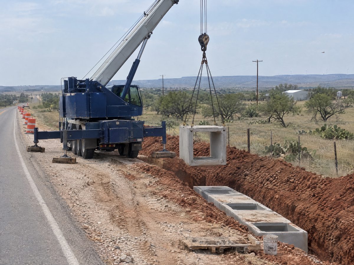 A dark blue truck crane on the shoulder of a two-lane highway lowers a precast concrete box culvert section on chain slings into a fresh ditch.